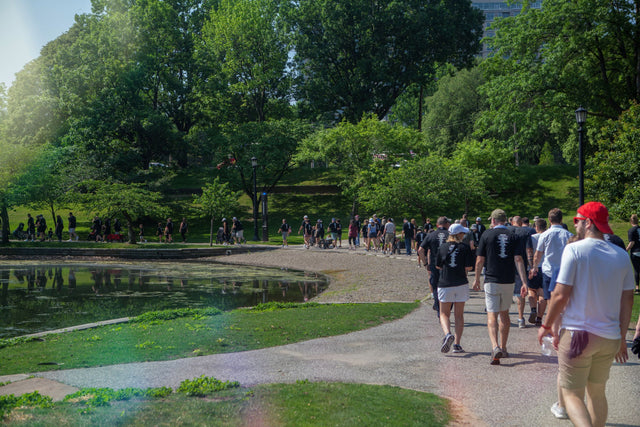 A group of people walk around Wade Lagoon in University Circle during White Cane Walk. The trees are leafy and green, and it's a bright sunny day.